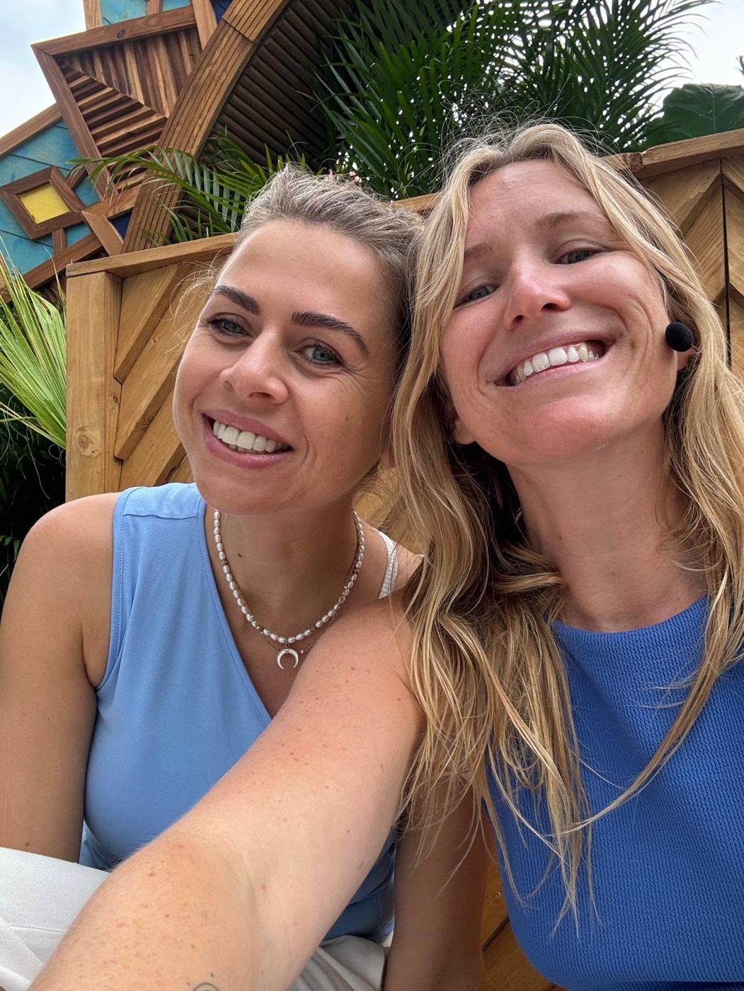 Cindy en Lydia lachend naast elkaar buiten voor een houten wand met palmbladeren